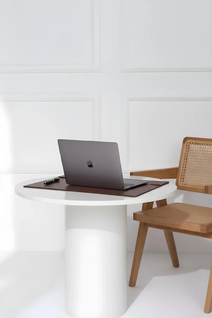 Na tle sztukateryjnej białej ściany stoi biały stolik a na nim czarny laptop. Obok stoi drewniane krzesło, jakby rattanowe.